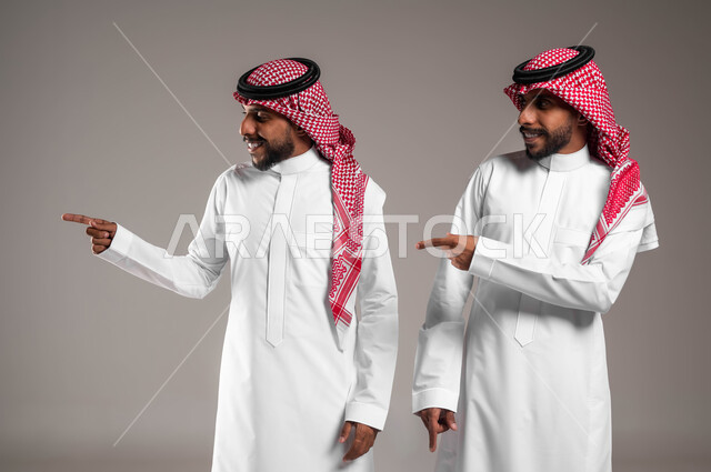 تعابير يدين تشير الى موقع معين, حركات تدل على هذا المكان (هنا), مفهوم لغة الجسد والإشارات، بورتريه لشابان توءم عربيان خليجيان سعوديان مبتسمان يرتديان الزي التقليدي ويشيران بأصابع السبابة جانبا إلى شيء ما بإيماءات السرور، خلفية رمادية .