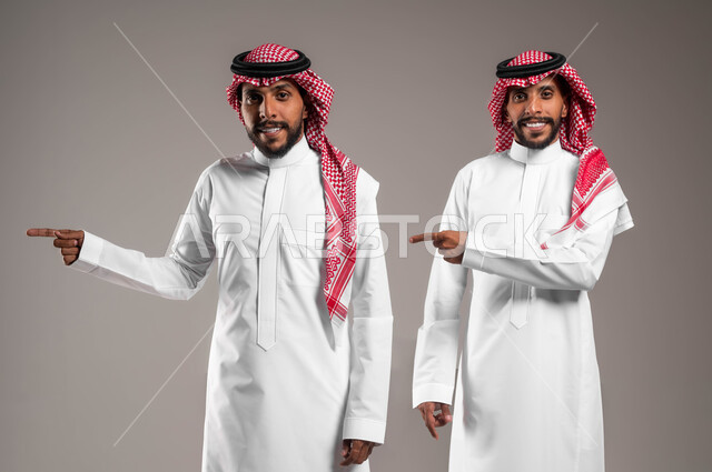 النظر الى الكاميرا بتعابير يدين تشير الى موقع معين, حركات تدل على هذا المكان (هنا), مفهوم لغة الجسد والإشارات، بورتريه لشابان توءم عربيان خليجيان سعوديان مبتسمان يرتديان الزي التقليدي ويشيران بأصابع السبابة جانبا إلى شيء ما بإيماءات السرور، خلفية رمادية