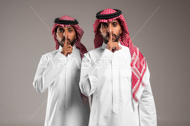 النظر إلى الكاميرا بتعابير طلب الهدوء وعدم الازعاج ،السكوت و التوقف عن الكلام, بورتريه مقرب لشابان توءم عربيان سعوديان خليجيان يرتديان الزي التقليدي يرفعان اصبعهما بإيماءات الامر بالصمت, الاشارة الى خفض الصوت والهمس, خلفية رمادية