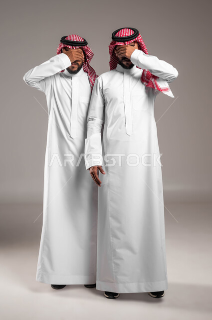 حركات تدل على اللعب و المرح, ايماءات الخجل و الاستعداد لرؤية المفاجأة, بورتريه لشابان توءم عربيان سعوديان خليجيان يرتديان الزي التقليدي ويضعان أيديهم على أعينهم, صورة الجسم بالطول الكامل، خلفية رمادية