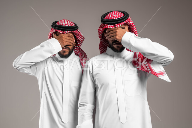 ايماءات الخجل و الاستعداد لرؤية المفاجأة, بورتريه مقرب لشابان توءم عربيان سعوديان خليجيان يرتديان الزي التقليدي ويضعان أيديهم على أعينهم, حركات تدل على اللعب و المرح, خلفية رمادية