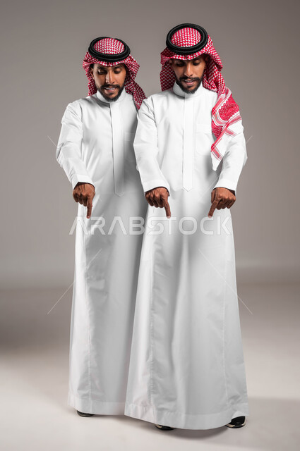 تعابير يدين تشير الى موقع معين, حركات تدل على هذا المكان (هنا), مفهوم لغة الجسد والإشارات، بورتريه لشابان توءم عربيان خليجيان سعوديان يرتديان الزي التقليدي ويشيران بأصابع السبابة نحو الاسفل إلى شيء ما بإيماءات السرور ،صورة الجسم بالطول الكامل، خلفية رمادية