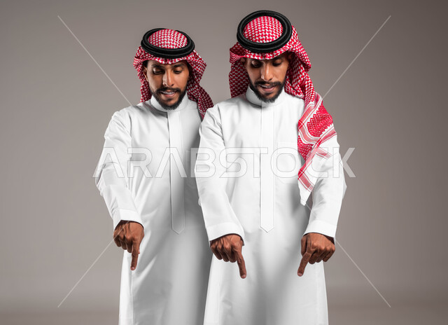 حركات تدل على هذا المكان (هنا), تعابير يدين تشير الى موقع معين, مفهوم لغة الجسد والإشارات، بورتريه لشابان توءم عربيان خليجيان سعوديان يرتديان الزي التقليدي ويشيران بأصابع السبابة نحو الاسفل إلى شيء ما ، خلفية رمادية