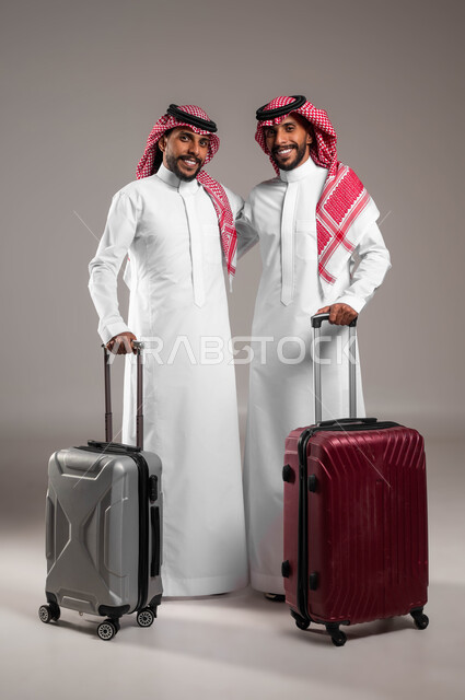 Looking at the camera with happy gestures, preparing to set off and start the tourist trip, spending an enjoyable summer vacation, the concept of leisure and entertainment, a portrait of two young Saudi Gulf Arab twins, smiling in traditional dress, holding a suitcase, full-length body portrait, gray background.