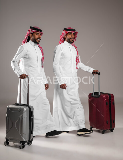 الاستعداد للانطلاق و بدء الرحلة السياحية، ايماءات المشي والتوجه نحو المطار، قضاء اجازة صيفية ممتعه, مفهوم الترفيه و التسلية, بورتريه لشابين توءم عربيين خليجيين سعوديين مبتسمين يرتديان الزي التقليدي يمسكان حقيبة السفر، صورة الجسم بالطول الكامل, خلفية رمادية .