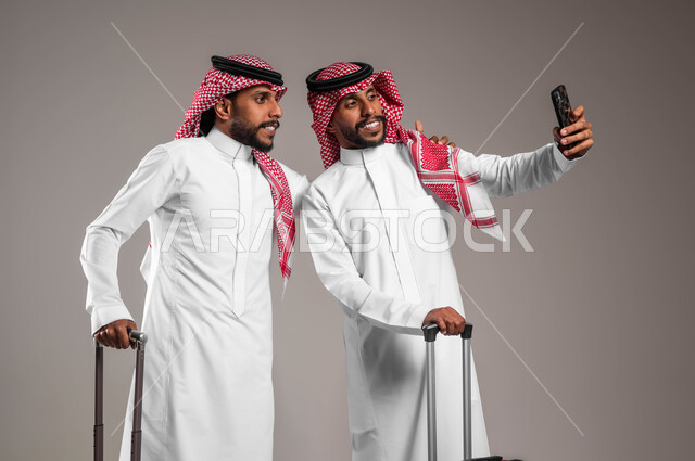 Using advanced modern technologies, preparing for an enjoyable tourist trip during the summer vacation, a portrait of two young Saudi Gulf Arab twins wearing traditional clothing, holding travel bags, documenting happy moments by taking souvenir selfies on a mobile phone, gray background.
