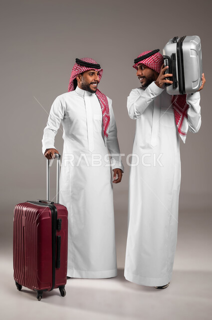 تبادل الأحاديث بتعابير السرور، قضاء العطلة الصيفيه خارج البلاد, بورتريه لشاب عربي خليجي سعودي يرتدي الثوب التقليدي و يحمل حقيبة السفر برفقة أخاه التوءم يستعدان لرحلة سياحيه ممتعه، مفهوم الترابط الأخوي، صورة الجسم بالطول الكامل، خلفية رمادية .