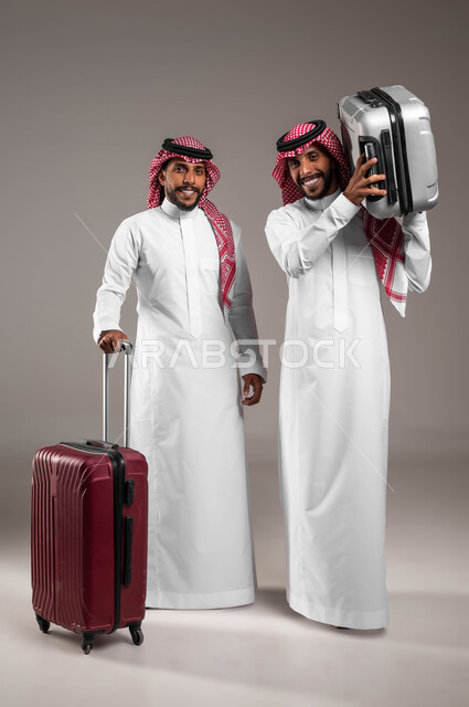 مفهوم الترابط الأخوي، النظر إلى الكاميرا بايماءات السرور، قضاء العطلة الصيفيه خارج البلاد, بورتريه لشاب عربي خليجي سعودي يرتدي الثوب التقليدي و يحمل حقيبة السفر برفقة أخاه التوءم يستعدان لرحلة سياحيه ممتعه، صورة الجسم بالطول الكامل، خلفية رمادية .