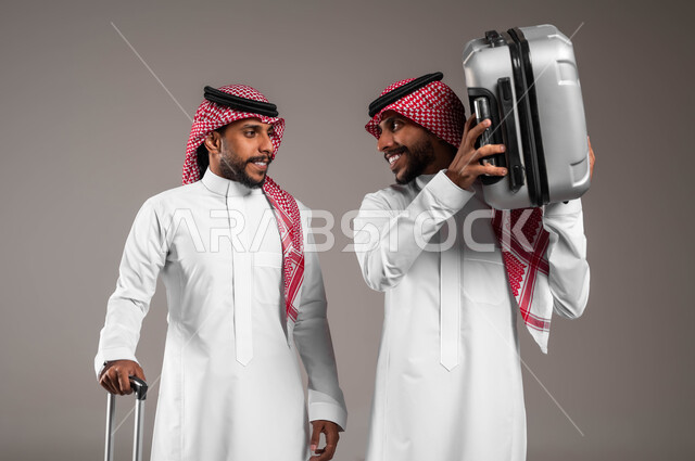 مفهوم الترفيه والتسلية بالعطلة الصيفية، قضاء أوقات الفراغ خارج البلاد, بورتريه لشاب عربي خليجي سعودي مبتسم يرتدي الثوب التقليدي و يحمل حقيبة السفر برفقة أخاه التوءم يستعدان لرحلة سياحيه ممتعه بتعابير السرور، مفهوم الترابط الأخوي، خلفية رمادية .