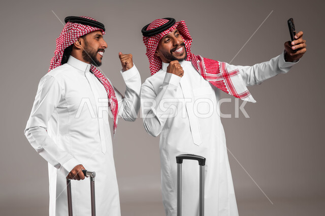 مفهوم السياحة و السفر, الاستعداد لقضاء رحلة ممتعة في إجازة الصيف، بورتريه لشابان توءم عربيان خليجيان سعوديان يرتديان الزي التقليدي ويقومان بتوثيق اللحظات السعيدة بالتقاط صور السيلفي التذكارية عبر الجوال، رفع الأيدي بعلامات النجاح والفوز ،خلفية رمادية