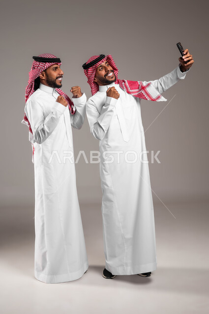استخدام التقنيات الحديثة المتطورة، توثيق اللحظات السعيدة، بورتريه لشاب عربي خليجي سعودي يرتدي الزي التقليدي و يقوم بالتقاط صور السيلفي التذكارية عبر الهاتف المحمول مع أخاه التوءم بإيماءات السرور، رفع اليدين بعلامات الفوز والانتصار، صورة لكامل الجسم، خلفية رمادية