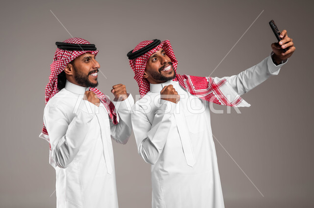 رفع اليدين بعلامات الفوز والانتصار، استخدام التقنيات الحديثة المتطورة، توثيق اللحظات السعيدة، بورتريه لشاب عربي خليجي سعودي يرتدي الزي التقليدي و يقوم بالتقاط صور السيلفي التذكارية عبر الهاتف المحمول مع أخاه التوءم بإيماءات السعادة والسرور، خلفية رمادية
