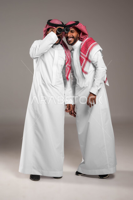 Future ambitions and goals, searching for something using binoculars, the concept of participating in exploration and adventure, a portrait of two young Saudi Gulf Arab twins wearing traditional clothing and sharing a view through binoculars, a full-body portrait, gray background.
