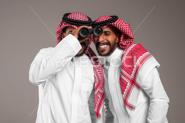 البحث عن شيء ما باستخدام الناظور، طموحات وأهداف مستقبلية، مفهوم المشاركة في الاستكشاف والمغامرة، بورتريه مقرب لشابان توءم عربيان خليجيان سعوديان يرتديان الزي التقليدي و يتشاركان في النظرعبر المنظار ثنائي العينين، خلفية رمادية .