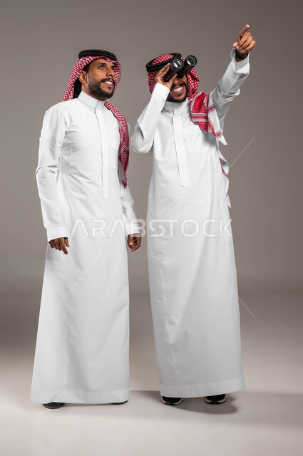 الأهداف والطموحات المستقبلية, مفهوم الاستكشاف والمغامرة, رؤية الأشياء عن قرب, بورتريه لشباب توءم عربيان خليجيان سعوديان يرتديان الثوب التقليدي والشماغ أحدهما ينظر إلى مكان معين والآخر يمسك المنظار ثنائي العينين, صورة الجسم بالطول الكامل, خلفية رمادية