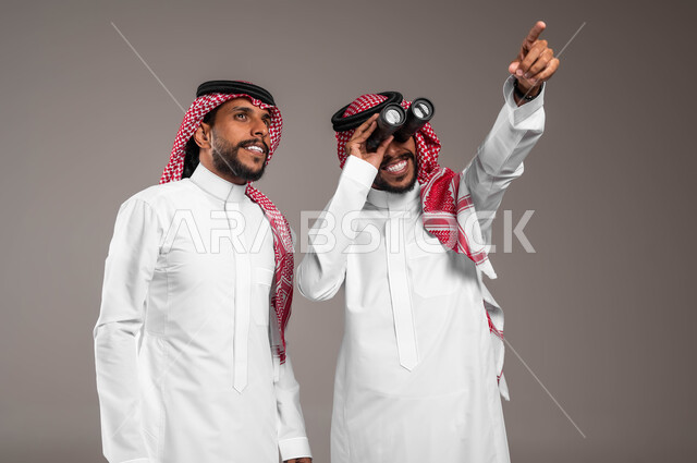 مفهوم الاستكشاف والمغامرة, الأهداف والطموحات المستقبلية, رؤية الأشياء عن قرب, بورتريه لشباب توءم عربيان خليجيان سعوديان يرتديان الثوب التقليدي والشماغ أحدهما ينظر إلى مكان معين والآخر يمسك المنظار ثنائي العينين, صورة الجسم بالطول الكامل, خلفية رمادية