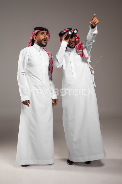 الطموحات والأهداف المستقبلية, مفهوم الاستكشاف والمغامرة, رؤية الأشياء عن قرب, بورتريه لشباب توءم عربيان خليجيان سعوديان يرتديان الثوب التقليدي والشماغ أحدهما ينظر إلى مكان معين والآخر يمسك المنظار ثنائي العينين, صورة الجسم بالطول الكامل, خلفية رمادية