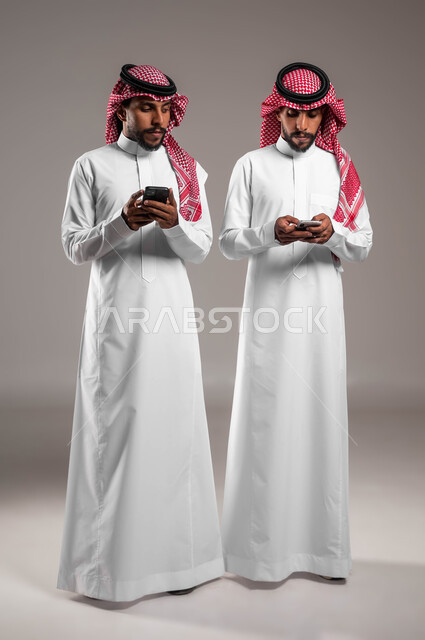 التطفل والتلصص على الهواتف الشخصية, اسخدام البرامج والتطبيقات الالكترونية, النظر خلسة للجوال الآخر, بورتريه لشباب توءم عربيان خليجيان سعوديان يرتديان الثوب التقليدي والشماغ يقومان باستخدام الهاتف المحمول, صورة الجسم بالطول الكامل, خلفية رمادية