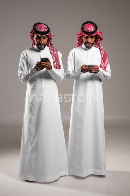 استخدام التطبيقات الالكترونية الحديثة, بورتريه لشباب توءم عربيان خليجيان سعوديان يرتديان الثوب التقليدي والشماغ يستخدمان الهاتف المحمول, التواصل مع الأصدقاء عبر وسائل السوشال ميديا, صورة الجسم بالطول الكامل, خلفية رمادية