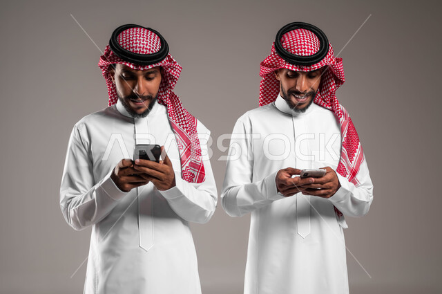 التواصل مع الأصدقاء عبر وسائل السوشال ميديا, استخدام التطبيقات الالكترونية الحديثة, بورتريه مقرب لشباب توءم عربيان خليجيان سعوديان يرتديان الثوب التقليدي والشماغ يستخدمان الهاتف المحمول, خلفية رمادية