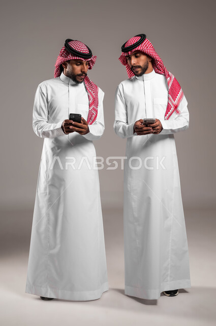 التلصص على الهواتف الشخصية, اسخدام البرامج والتطبيقات الالكترونية, التطفل والنظر خلسة للجوال الآخر, بورتريه لشباب توءم عربيان خليجيان سعوديان يرتديان الثوب التقليدي والشماغ يقومان باستخدام الهاتف المحمول, صورة الجسم بالطول الكامل, خلفية رمادية