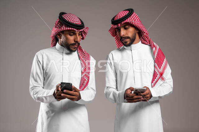 اسخدام البرامج والتطبيقات الالكترونية, التطفل والنظر خلسة للجوال الآخر, بورتريه مقرب لشباب توءم عربيان خليجيان سعوديان يرتديان الثوب التقليدي والشماغ يقومان باستخدام الهاتف المحمول, التلصص على الهواتف الشخصية, خلفية رمادية