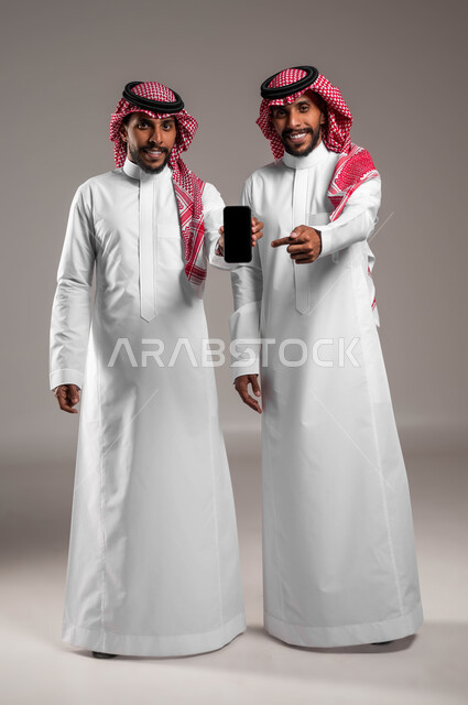 Pointing with a finger at a mobile phone with a blank black screen, a portrait of young Saudi Gulf Arab twins wearing the traditional dress and shemagh looking at the camera, using modern devices and technologies, full-length body photo, gray background