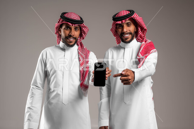 الوقوف و الإشارة الى الهاتف المحمول بشاشة سوداء فارغة, بورتريه لشباب توءم عربيان خليجيان سعوديان يرتديان الثوب التقليدي والشماغ ينظران إلى الكاميرا, استخدام الأجهزة والتقنيات الحديثة, خلفية رمادية