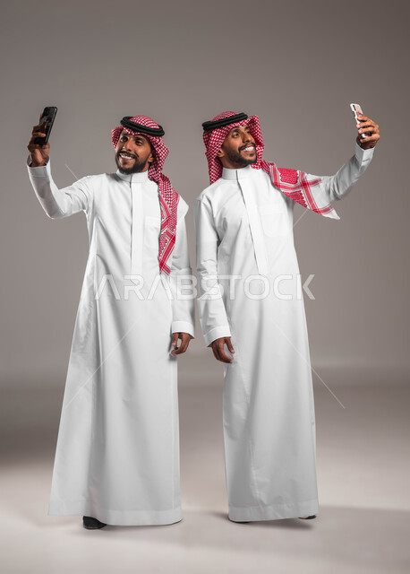 توثيق اللحظات السعيدة, الاستمتاع بالتقاط صور سيلفي تذكارية عبر الجوال, بورتريه لشباب توءم عربيان خليجيان سعوديان يرتديان الثوب التقليدي والشماغ يقومان باستخدام الهاتف المحمول, استخدام الأجهزة والتقنيات الحديثة, صورة الجسم بالطول الكامل, خلفية رمادية