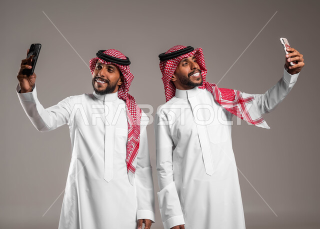الاستمتاع بالتقاط صور سيلفي تذكارية عبر الجوال, بورتريه لشباب توءم عربيان خليجيان سعوديان يرتديان الثوب التقليدي والشماغ يقومان باستخدام الهاتف المحمول, توثيق اللحظات السعيدة, استخدام الأجهزة والتقنيات الحديثة, خلفية رمادية