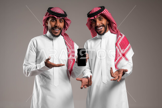استخدام الأجهزة والتقنيات الحديثة, النظر إلى الكاميرا بإيماءات الدهشة والسؤال والاستغراب, بورتريه مقرب لشباب توءم عربيان خليجيان سعوديان يرتديان الثوب التقليدي والشماغ أحدهما يمسك الهاتف المحمول بشاشة سوداء فارغة, خلفية رمادية