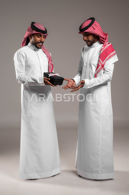 الوقوف باستقامة بإيماءات السرور, التعبير عن المحبة والألفة بين الأخوان, أعياد ومناسبات ومفاجآت سعيدة, تقديم الهدايا التذكارية, بورتريه لشاب عربي خليجي سعودي يرتدي الثوب التقليدي والشماغ يقوم بتقديم الهدية لأخيه التوءم, صورة الجسم بالطول الكامل, خلفية رمادية