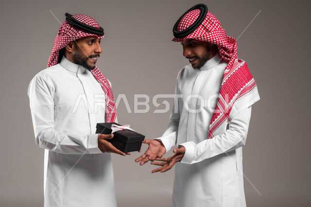 أعياد ومناسبات ومفاجآت سعيدة, تقديم الهدايا التذكارية, التعبير عن المحبة والألفة بين الأخوان, بورتريه مقرب لشاب عربي خليجي سعودي يرتدي الثوب التقليدي والشماغ يقوم بتقديم الهدية لأخيه التوءم, خلفية رمادية