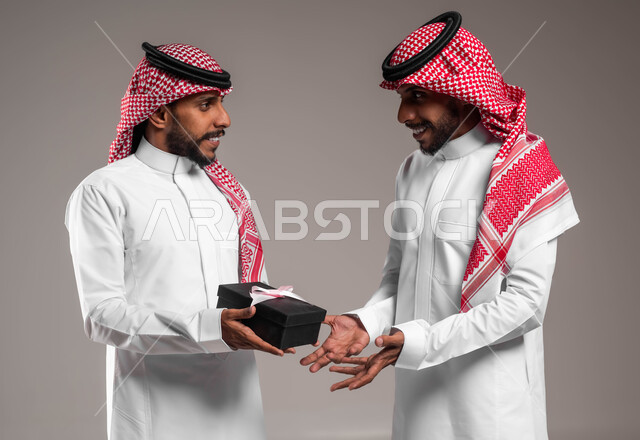 تقديم الهدايا التذكارية, التعبير عن المحبة والألفة بين الأخوان, بورتريه مقرب لشاب عربي خليجي سعودي يرتدي الثوب التقليدي والشماغ يقوم بتقديم الهدية لأخيه التوءم, أعياد ومناسبات ومفاجآت سعيدة, خلفية رمادية