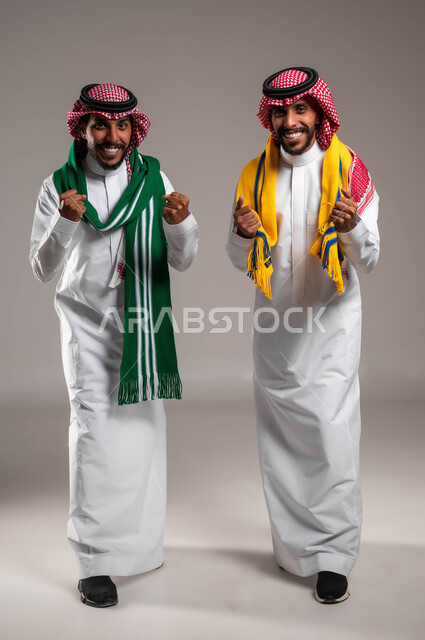 النظر إلى الكاميرا بتفاعل واندماج وحماس, البشاشة والسرور لتحقيق الفوز والانتصار, الشغف بمتابعة المباريات الرياضية, بورتريه لشباب توءم عربيان خليجيان سعوديان يرتديان الثوب التقليدي والشماغ يرتديان أوشحة تشجيعية, صورة الجسم بالطول الكامل, خلفية رمادية