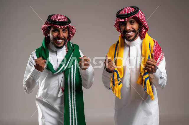 التفاعل والاندماج والحماس بمشاهدة الألعاب الرياضية, البشاشة والسرور لتحقيق الفوز والانتصار, الشغف بمتابعة المباريات, بورتريه مقرب لشباب توءم عربيان خليجيان سعوديان يرتديان الثوب التقليدي والشماغ يرتديان أوشحة تشجيعية, خلفية رمادية