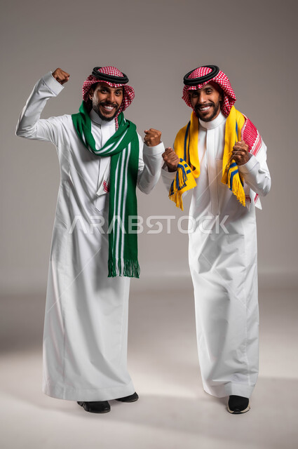 البشاشة والسرور لتحقيق الفوز والانتصار, النظر إلى الكاميرا بتفاعل واندماج وحماس, الشغف بمتابعة المباريات الرياضية, بورتريه لشباب توءم عربيان خليجيان سعوديان يرتديان الثوب التقليدي والشماغ يرتديان أوشحة تشجيعية, صورة الجسم بالطول الكامل, خلفية رمادية