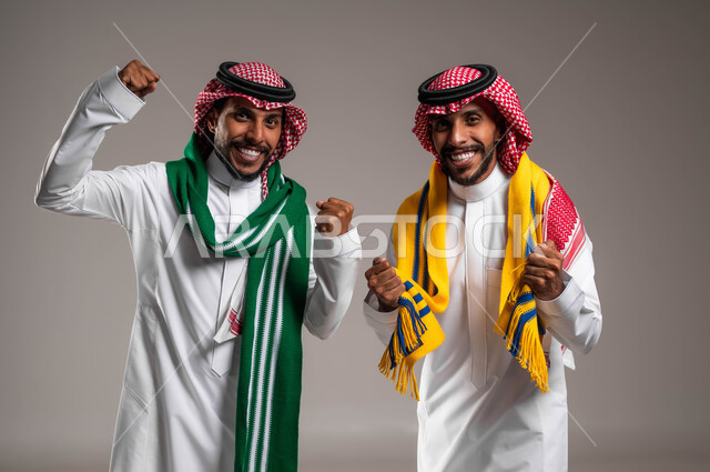 الاستمتاع والشغف بمتابعة المباريات الرياضية, السرور والبشاشة لتحقيق الفوز والانتصار, النظر إلى الكاميرا بتفاعل واندماج وحماس, بورتريه مقرب لشباب توءم عربيان خليجيان سعوديان يرتديان الثوب التقليدي والشماغ يرتديان أوشحة تشجيعية, خلفية رمادية
