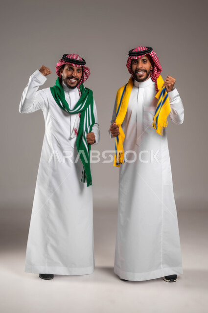 الشغف بمتابعة المباريات الرياضية, السرور والبشاشة لتحقيق الفوز والانتصار, النظر إلى الكاميرا بتفاعل واندماج وحماس, بورتريه لشباب توءم عربيان خليجيان سعوديان يرتديان الثوب التقليدي والشماغ يرتديان أوشحة تشجيعية, صورة الجسم بالطول الكامل, خلفية رمادية