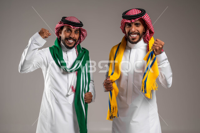 Happiness and joy to achieve victory and triumph, looking at the camera with interaction, integration and enthusiasm, passion for following sports matches, portrait of young Saudi Gulf Arab twins wearing traditional thobes and shemaghs, wearing encouragement scarves, gray background
