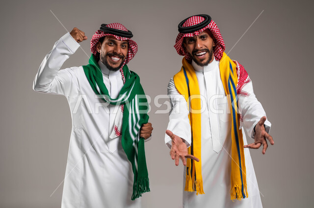 النظر إلى الكاميرا بتفاعل واندماج وحماس, الشغف بمتابعة المباريات الرياضية, السرور والبشاشة لتحقيق الفوز والانتصار, بورتريه مقرب لشباب توءم عربيان خليجيان سعوديان يرتديان الثوب التقليدي والشماغ يرتديان أوشحة تشجيعية, خلفية رمادية