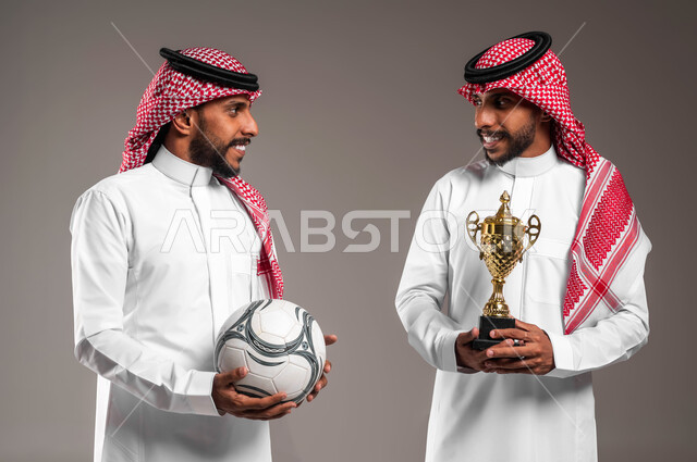 الوقوف بإيماءات السرور, تحقيق الفوز والانتصار في المباراة, بورتريه مقرب لشباب توءم عربيان خليجيان سعوديان يرتديان الثوب التقليدي والشماغ أحدهما يحمل كأس البطولة والآخر كرة القدم, متابعة المبارايات والأنشطة الرياضية, خلفية رمادية