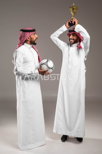 المنافسة والتحديات الرياضية, التعبير بحماس وسرور لتحقيق الفوز والانتصار, بورتريه لشباب توءم عربيان خليجيان سعوديان يرتديان الثوب التقليدي والشماغ أحدهما يرفع كأس البطولة على رأسه والآخر يحمل كرة القدم, الشغف بمتابعة المباراة, صورة الجسم بالكامل, خلفية رمادية