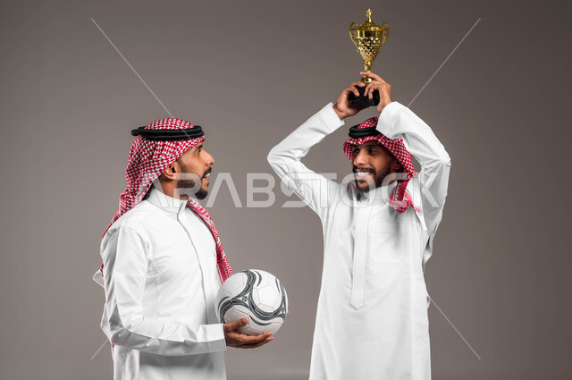 التعبير بحماس وسرور لتحقيق الفوز والانتصار, بورتريه مقرب لشباب توءم عربيان خليجيان سعوديان يرتديان الثوب التقليدي والشماغ أحدهما يرفع كأس البطولة على رأسه والآخر يحمل كرة القدم, الشغف بمتابعة المباراة, المنافسة والتحديات الرياضية, خلفية رمادية