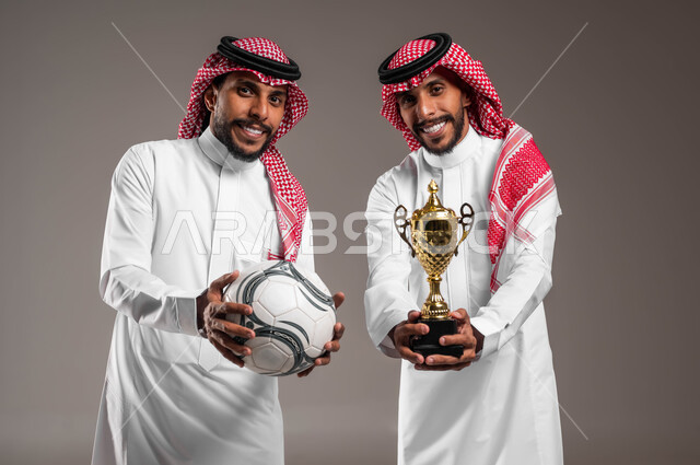 الوقوف بسرور والنظر إلى الكاميرا, بورتريه مقرب لشباب توءم عربيان خليجيان سعوديان يرتديان الثوب التقليدي والشماغ أحدهما يحمل كأس البطولة والآخر يمسك كرة القدم, الانتصار والفوز في المباراة, أنشطة رياضية ترفيهية, خلفية رمادية