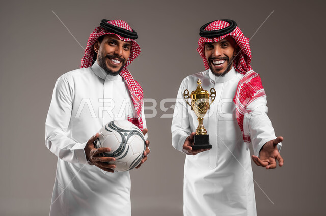 أنشطة رياضية ترفيهية, الفوز والانتصار في المباراة, التشجيع والشغف بمتابعة المباريات الرياضية, بورتريه مقرب لشباب توءم عربيان خليجيان سعوديان يرتديان الثوب التقليدي والشماغ أحدهما يمسك كأس البطولة والآخر يحمل الكرة بإيماءات البشاشة والسرور, خلفية رمادية