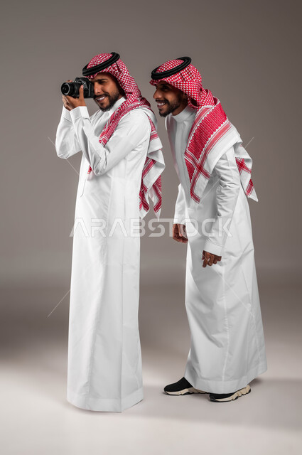 توثيق اللحظات السعيدة, استخدام كاميرا فوتوغرافية عالية الدقة, الاستمتاع بممارسة هواية فن التصوير, بورتريه لتوءم شباب عربيين خليجين سعوديين يرتديان الثوب التقليدي والشماغ أحدهما يمسك الكاميرا والآخر ينظر إلى شيء ما, صورة الجسم بالكامل, خلفية رمادية