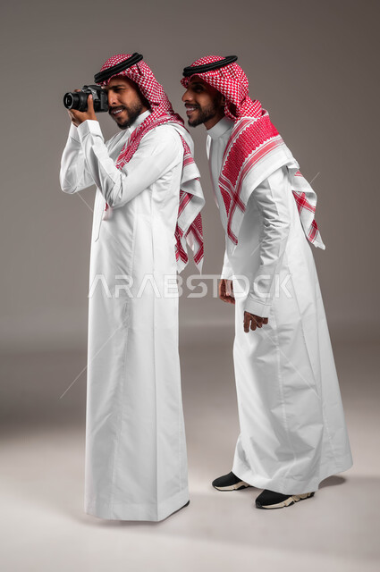 استخدام كاميرا فوتوغرافية عالية الدقة, الاستمتاع بممارسة هواية فن التصوير, بورتريه لتوءم شباب عربيين خليجين سعوديين يرتديان الثوب التقليدي والشماغ أحدهما يمسك الكاميرا والآخر ينظر إلى شيء ما, توثيق اللحظات السعيدة, صورة الجسم بالكامل, خلفية رمادية