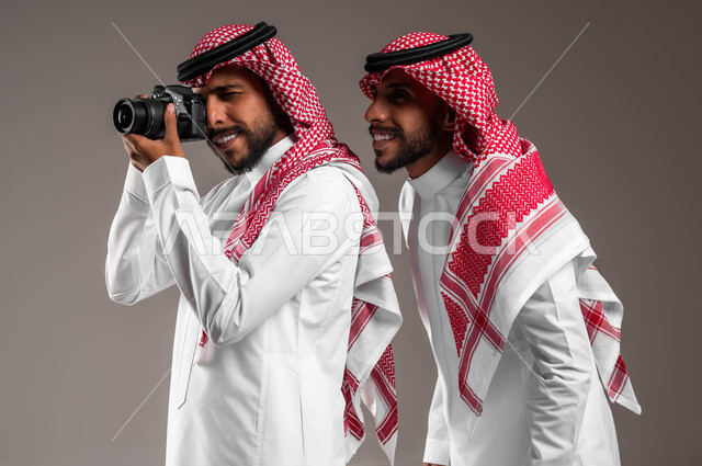 الاستمتاع بممارسة هواية فن التصوير, استخدام كاميرا فوتوغرافية عالية الدقة, بورتريه مقرب لتوءم شباب عربيين خليجين سعوديين يرتديان الثوب التقليدي والشماغ أحدهما يمسك الكاميرا والآخر ينظر إلى شيء ما, توثيق اللحظات السعيدة, خلفية رمادية