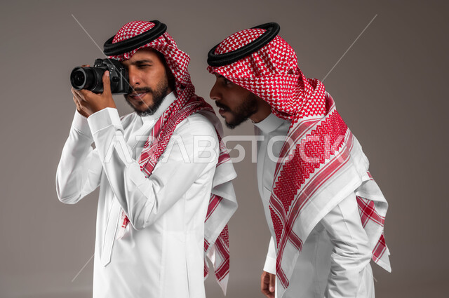 Scrutinizing and focusing on taking pictures, documenting happy moments, a close-up portrait of twin Saudi Gulf Arab men wearing the traditional dress and shemagh, using a high-resolution camera, professional photography in Saudi Arabia, gray background